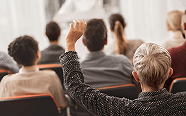 Image of many people sitting at a meeting with hands help up waiting to be chosen to speak.
