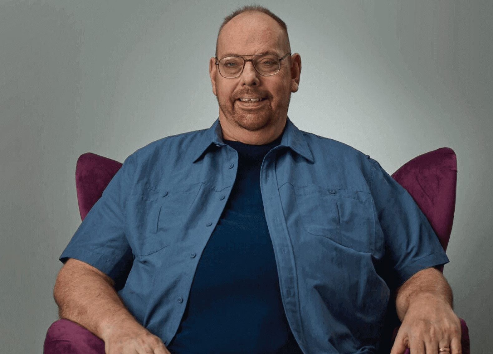 Image of a man in a blue shirt sitting in a purple chair.