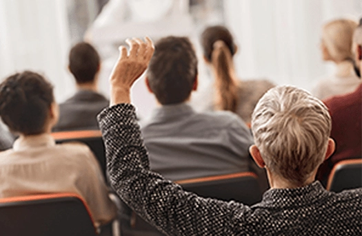 Image of many people sitting at a meeting with hands held up waiting to be chosen to speak.